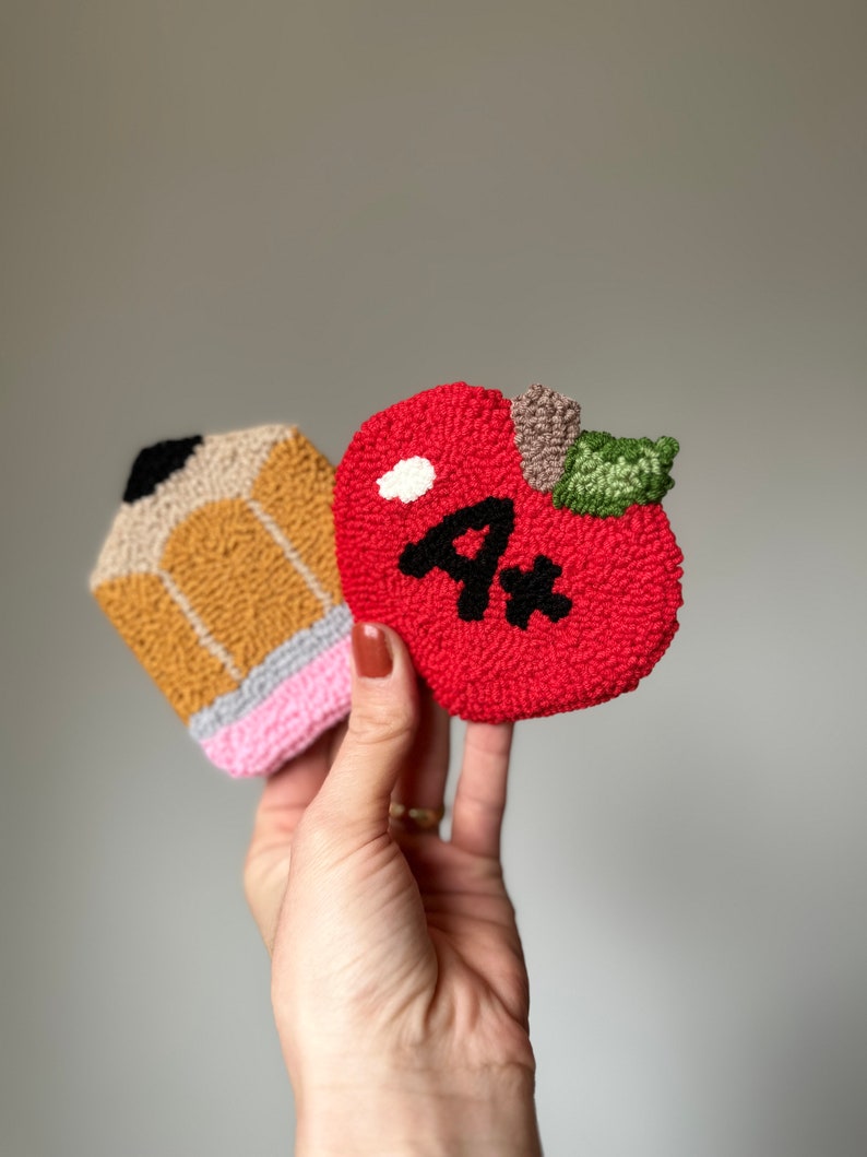May include: A red felt apple with a green stem and a black "A+" on it. A yellow and brown felt pencil with a pink eraser is next to the apple.