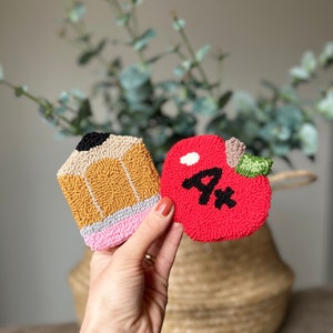May include: A red felt apple with a white stem and a black "A+" on it. A yellow and brown felt pencil with a pink eraser is next to the apple.
