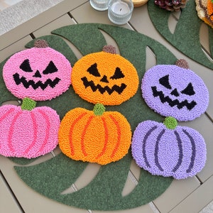 May include: Six handmade pumpkin-shaped coasters with different colors and designs. The coasters are made of yarn and have a fluffy texture. They are arranged on a green leaf-shaped mat.