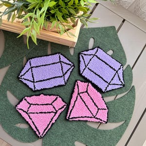 May include: Four handmade coasters shaped like purple and pink gemstones. The coasters are made with a looped yarn technique and have black outlines.