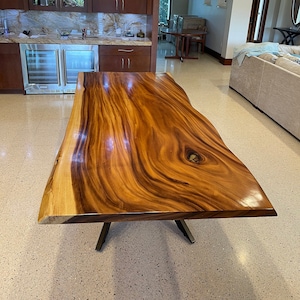 May include: A large, live edge wooden table with a natural wood finish. The table top has a unique grain pattern and a knot in the center. The table is supported by a simple, black metal base.