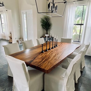 May include: A large, live edge wood dining table with a natural wood finish. The table is surrounded by white slipcovered chairs. A black geometric chandelier hangs above the table.