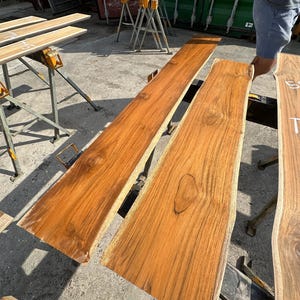 May include: Two large pieces of wood, likely teak, with a natural, live edge finish. The wood is a rich brown color with a warm, golden hue. The wood is sitting on a concrete surface.