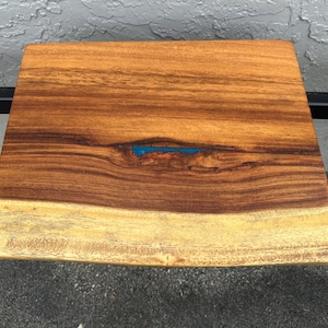 May include: A square wooden cutting board with a natural edge. The board is made of dark brown wood with a lighter, yellowish edge. A small, rectangular inlay of blue resin is visible in the center.