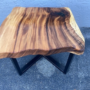 May include: A square wooden coffee table with a live edge top and a black metal base. The table has a natural wood finish and a rustic look.
