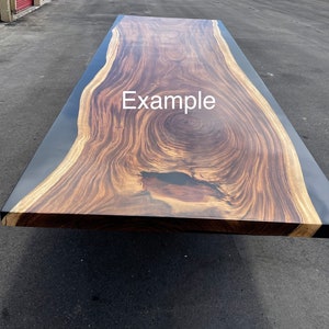 May include: A large, dark wood table top with a live edge and black epoxy resin filling. The table top has a natural wood grain pattern and a large knot in the center. The text "Example" is written on the table top.
