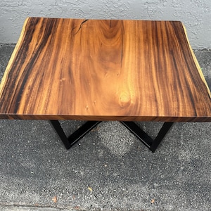 May include: A wooden table top with a live edge and a natural wood finish. The table top is supported by a black metal base with a criss-cross design.