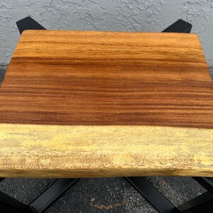 May include: A wooden table top with a natural edge and a dark brown and light yellow wood grain pattern. The table top is supported by a black metal base.