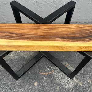 May include: A wooden bench with a natural wood finish and a black metal X-shaped base.