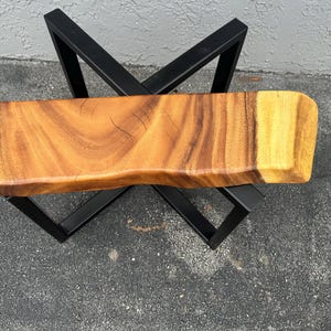 May include: A wooden table top with a live edge and a natural wood finish. The table top is supported by a black metal X-shaped base.