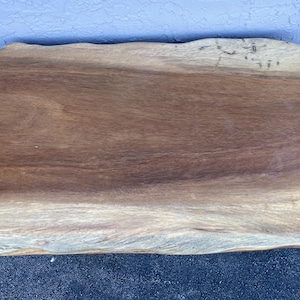 May include: A large, natural wood slab table top with a live edge. The wood is a rich brown color with a natural grain pattern. The table top is supported by a black metal base.