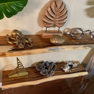 May include: Two wooden shelves with a live edge design. The top shelf has a gold octopus figurine, a gold and white shell, and a silver metal sculpture. The bottom shelf has a gold sailboat figurine, a coral sculpture, and a gold fish figurine.