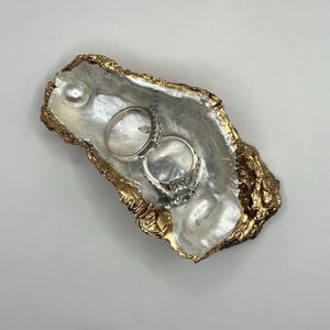 May include: A decorative oyster shell ring dish with a gold-colored trim. The dish contains two silver rings and a single pearl. The interior of the shell has a pearlescent sheen.