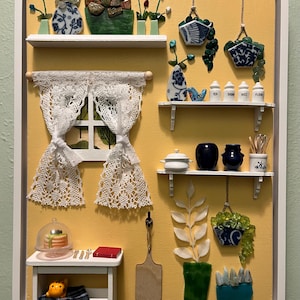 May include: A miniature kitchen scene with a yellow background and white frame. It features shelves with decorative items like plants, jars, and a miniature table with a cake. The scene includes a lace curtain and hanging planters, creating a charming, detailed display.
