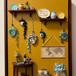 May include: A framed artwork featuring a mustard-yellow background with miniature decorative elements. It includes shelves with small dishes and vases, hanging planters, a table with a lamp and wine, a map, a basket, and a dog figurine.