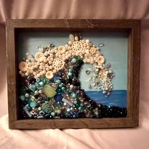 May include: A framed artwork depicting a wave, crafted from various beads, buttons, and embellishments. The wave crest is composed of white buttons and beads, transitioning to shades of blue and green. The artwork is set against a light blue background.