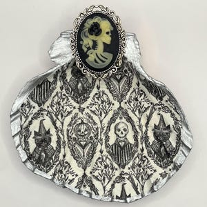 May include: A decorative Halloween-themed trinket dish. The dish is shaped like a seashell with a silver-painted rim. The interior features a black and white pattern with Halloween characters. A cameo with a skeleton design is attached.