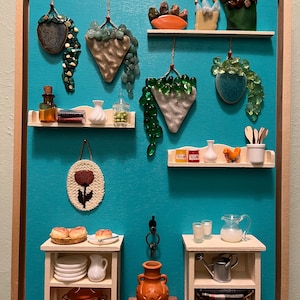 May include: A framed miniature kitchen scene with a teal background. It features small shelves with various items like miniature food, vases, and hanging decorative pieces. The scene includes a small table with plates and a pitcher, and another with a watering can.