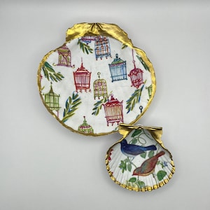 May include: Two decorative gold-rimmed seashells with a white background and a floral pattern. The larger shell has a pattern of birds and cages. The smaller shell has a pattern of birds and flowers.