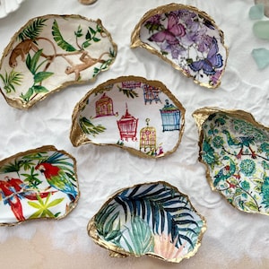 May include: Six decorative seashells with gold trim, each featuring a different patterned design. The designs include monkeys in a jungle, birds in a garden, tropical leaves, and cages with birds.