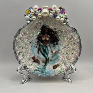 May include: A circular decorative dish featuring a mermaid illustration with dark skin and a teal tail, surrounded by iridescent glitter. The top edge is adorned with white pearls and faceted crystals, supported by a silver metal stand.