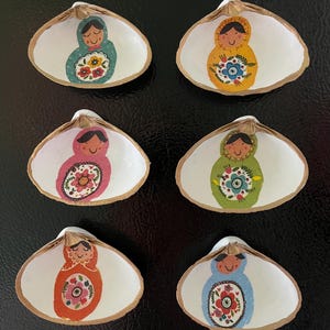 May include: Six painted seashell ornaments. Each shell is white inside with a painted figure wearing a hooded garment and holding a floral design. The garments are in various colors, including teal, yellow, pink, green, orange, and blue. The shells have a gold-colored trim.