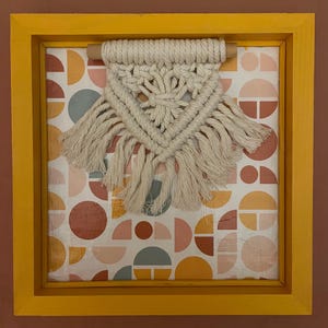 May include: A framed macrame wall hanging. The macrame is cream-colored and triangular, with a fringe at the bottom. The background features a geometric pattern of semi-circles in shades of orange, pink, and gray. The frame is a bright yellow.