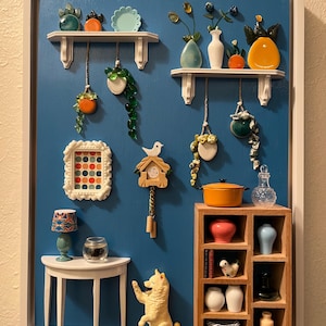 May include: A miniature diorama featuring a blue wall with white shelves and various decorative items. Includes small vases, hanging plants, a cuckoo clock, a table with a lamp, a dog figurine, and a wooden cabinet with miniature objects.