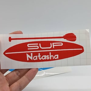 Pode incluir: Adesivo de vinil vermelho e branco com um design de remo e prancha de surf. O texto "SUP Natasha" está no adesivo.