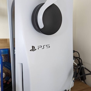 Pode incluir: Um console PlayStation 5 branco com um logotipo PlayStation preto e o texto "PS5" na frente. Um fone de ouvido sem fio preto está pendurado no console.