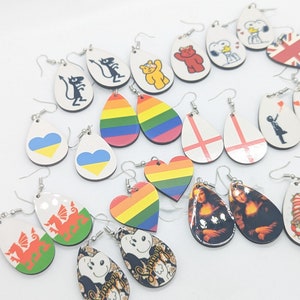 Puede incluir: Surtido de pendientes colgantes coloridos y únicos con varios diseños, incluyendo banderas, personajes de dibujos animados y patrones abstractos. Los pendientes están hechos de madera y tienen ganchos plateados.