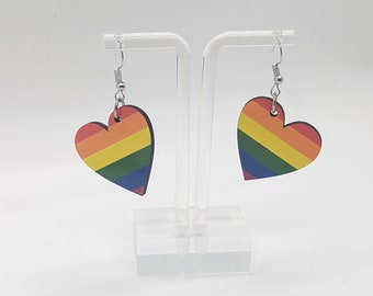 Könnte beinhalten: Zwei herzförmige Ohrringe mit Regenbogen-Pride-Flaggen-Design. Die Ohrringe hängen an silberfarbenen Fischhaken-Ohrdrähten. Sie werden auf einem transparenten Acryl-Ohrringständer vor weißem Hintergrund präsentiert.