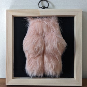 A framed piece of art featuring a pink fluffy object in a pine wooden frame with a black background.