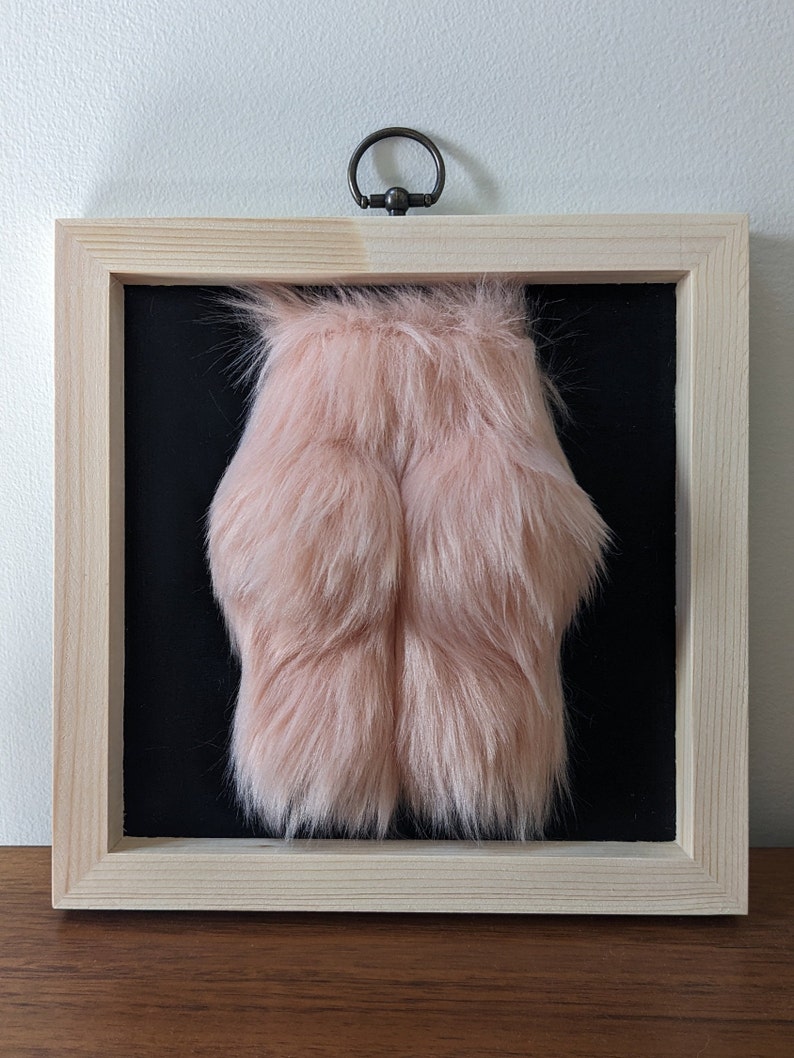 A framed piece of art featuring a pink fluffy object in a pine wooden frame with a black background.
