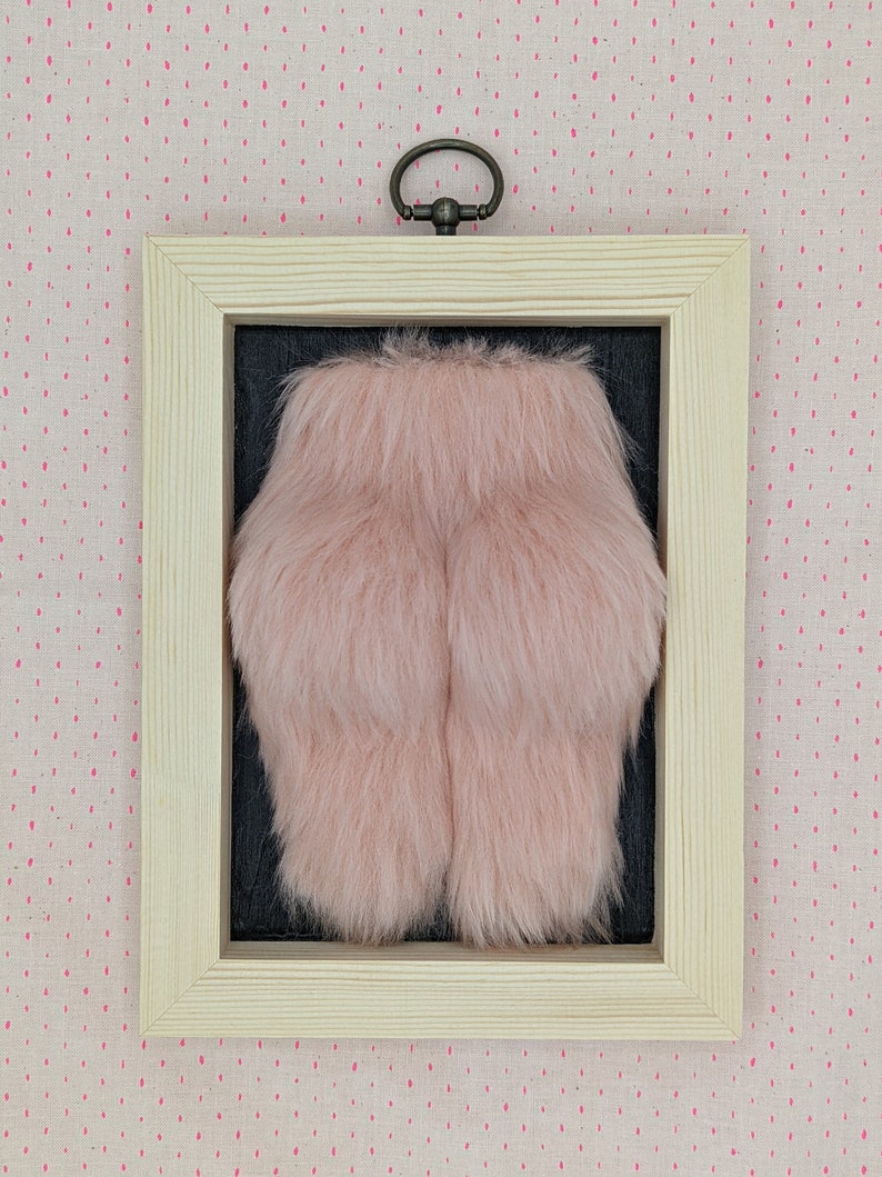 A framed piece of art featuring a pink fluffy object in a pine wooden frame with a black background.