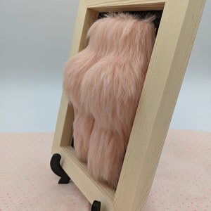 Side view of a framed piece of art featuring a pink fluffy object in a pine wooden frame with a black background.
