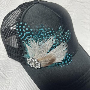 May include: A black trucker hat featuring a mesh back and a solid front panel. The front is decorated with a cluster of teal and black spotted feathers layered with light brown feathers, centered with a round silver-tone rhinestone brooch.