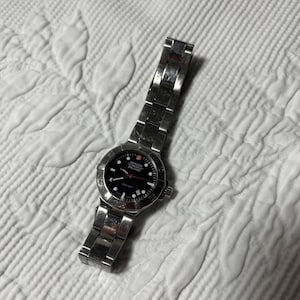 May include: A silver-toned stainless steel Wenger Swiss Military wristwatch featuring a round black dial with white hour markers, a red second hand, and a date window. The watch has a matching metal link bracelet and a rotating bezel.