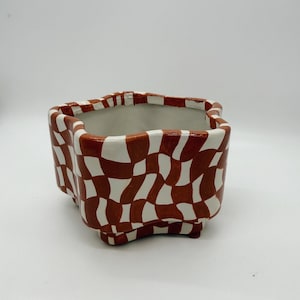 May include: A square ceramic planter with a brown and white checkered pattern. The planter has a unique shape with rounded edges and small feet. The interior is white, and the exterior features a repeating design. This decorative planter is suitable for indoor plants.