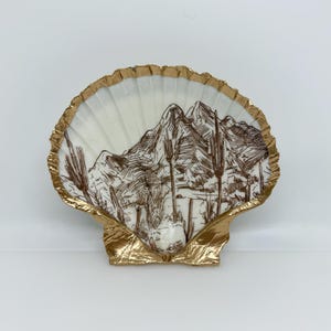 Könnte beinhalten: Eine dekorative Muschelschale mit goldfarbenem Rand. Die Schale zeigt eine braune Illustration einer Wüstenlandschaft mit Bergen und Kakteen. Die Schale ist weiß mit goldfarbener Verzierung.