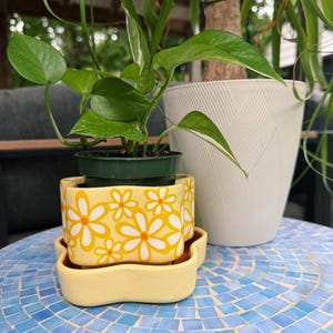 May include: A small potted plant sits atop a yellow daisy-patterned planter and saucer. A larger white planter with a textured design and a tall plant are in the background. The table has a blue mosaic tile design.