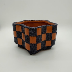 May include: A square-shaped ceramic planter with a black and orange checkerboard pattern. The planter has a textured, speckled exterior and a smooth, orange interior. The design is a modern take on a classic pattern, suitable for home decor.
