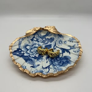 May include: A decorative seashell dish with a blue and white floral pattern and gold trim. Two gold hoop earrings rest inside the dish. The dish is a unique and elegant jewelry holder.