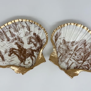 May include: Two decorative seashell dishes with a gold rim. The white interior features a brown illustration of cowboys on horseback and desert landscapes. A unique home decor item.