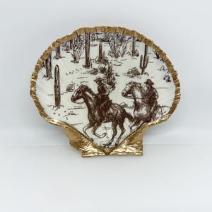 Op de afbeelding: Een decoratieve schelpvormige plaat met een goud geschilderde rand. De plaat heeft een sepia-getinte illustratie van cowboys die paardrijden door een woestijnlandschap met cactussen en andere woestijnplanten. De plaat staat op een witte achtergrond.