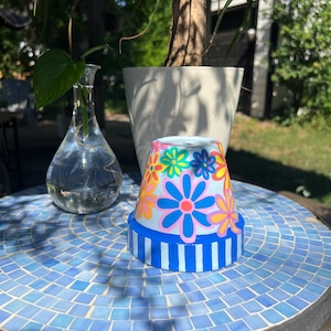 May include: A hand-painted flower pot with a blue and white striped base and colorful floral design. The pot sits on a mosaic tile table next to a glass vase with a plant cutting and a white planter.