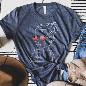 May include: A navy blue t-shirt with a white line drawing of a woman wearing sunglasses with an American flag design. The text "stars stripes girl power" is written below the woman's image.