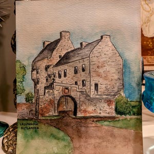 Può includere: Acquerello del castello di Lallybroch di Outlander. L'opera raffigura un castello in pietra con un ingresso ad arco, una strada marrone che conduce ad esso e un cielo blu. Il testo "Lallybroch Outlander" è visibile.