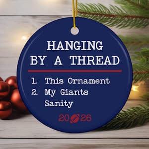 May include: A round, dark blue ceramic ornament with a gold hanging string. The text reads: HANGING BY A THREAD, 1. This Ornament, 2. My Giants Sanity, and 2026 with a football icon. A red horizontal line separates the title from the list.