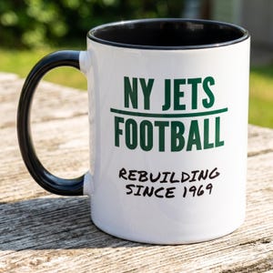May include: A white ceramic mug with a black handle and interior. The front features the green text NY JETS FOOTBALL above a horizontal line, with the black text REBUILDING SINCE 1969 printed below. The mug has a smooth, glossy finish.